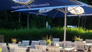 An outdoor seating area with tables and chairs under a large umbrella, surrounded by greenery. - BASALT Hotel Restaurant Lounge (Hohwacht)