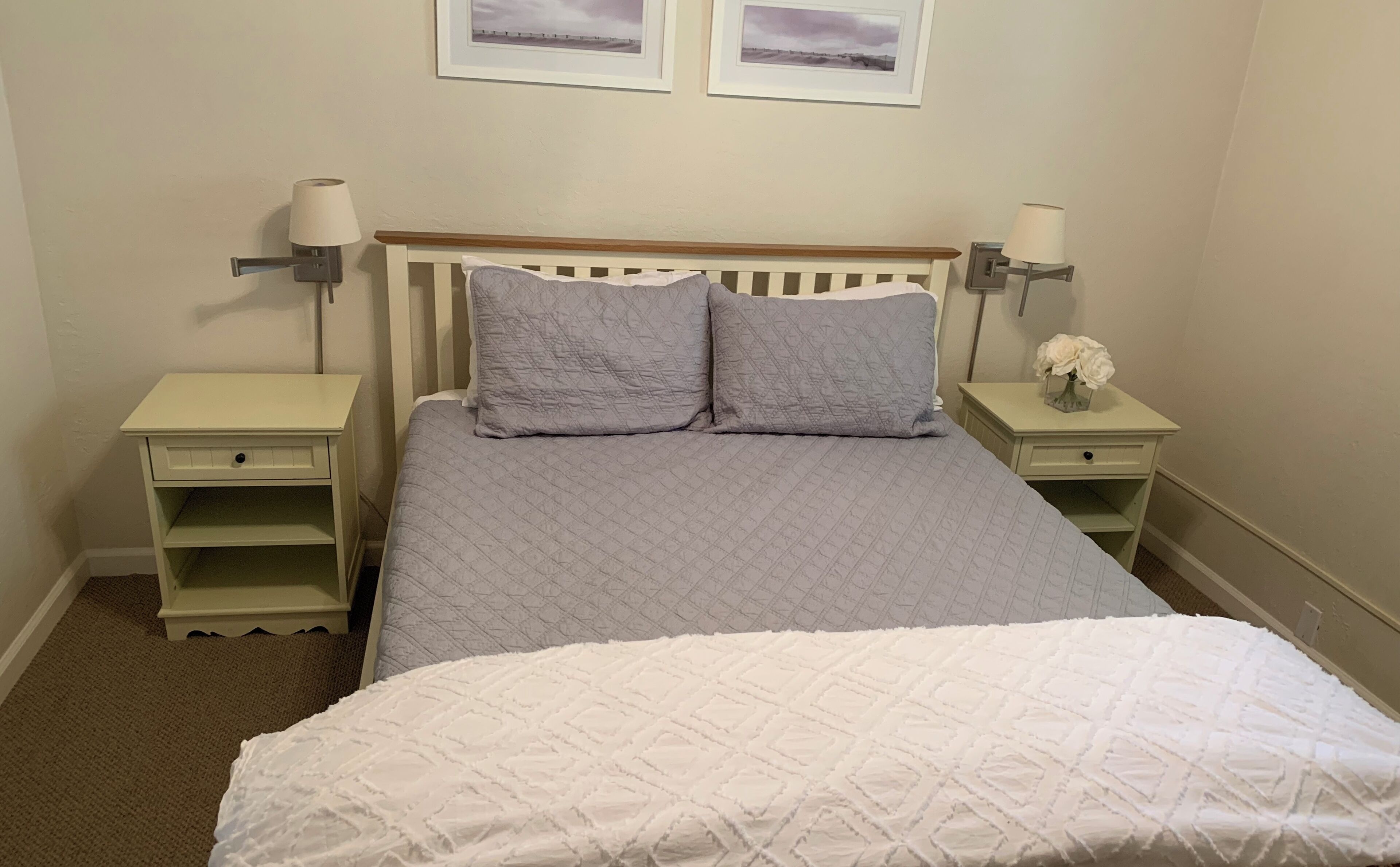 A neatly made bed with a white and gray quilt, two bedside tables with lamps, and a small vase with flowers on one table.