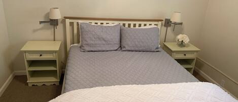 A neatly made bed with a white and gray quilt, two bedside tables with lamps, and a small vase with flowers on one table.