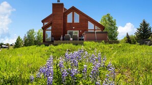 Een modern huis met een ruime voortuin, een houten terras en een helderblauwe lucht.
