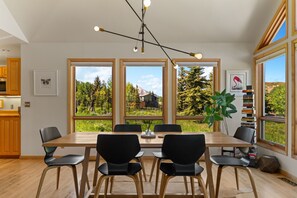 A modern dining area with a wooden table and black chairs, large windows with a view of greenery and a building, and a ceiling light fixture.
