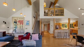 Een ruime woonkamer met een hoge plafond, houten balken, een trap naar een loft en een keuken met krukjes.
