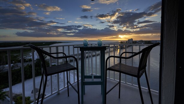 A balcony with two wicker chairs and a small table, overlooking a coastal view with a sunset.