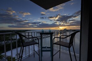 Ein Balkon mit zwei Korbstühlen und einem kleinen Tisch, der Blick auf die Küste und einen Sonnenuntergang bietet.