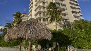 A tall hotel with multiple balconies, a beach area with lounge chairs, and a thatched umbrella.