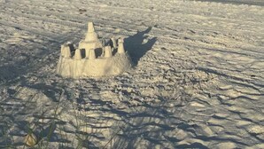 A sandcastle on a beach with a calm sea and distant buildings.