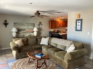 A living room with a sectional sofa, a coffee table, and a rug. There is a ceiling fan, a painting, and a kitchen in the background.