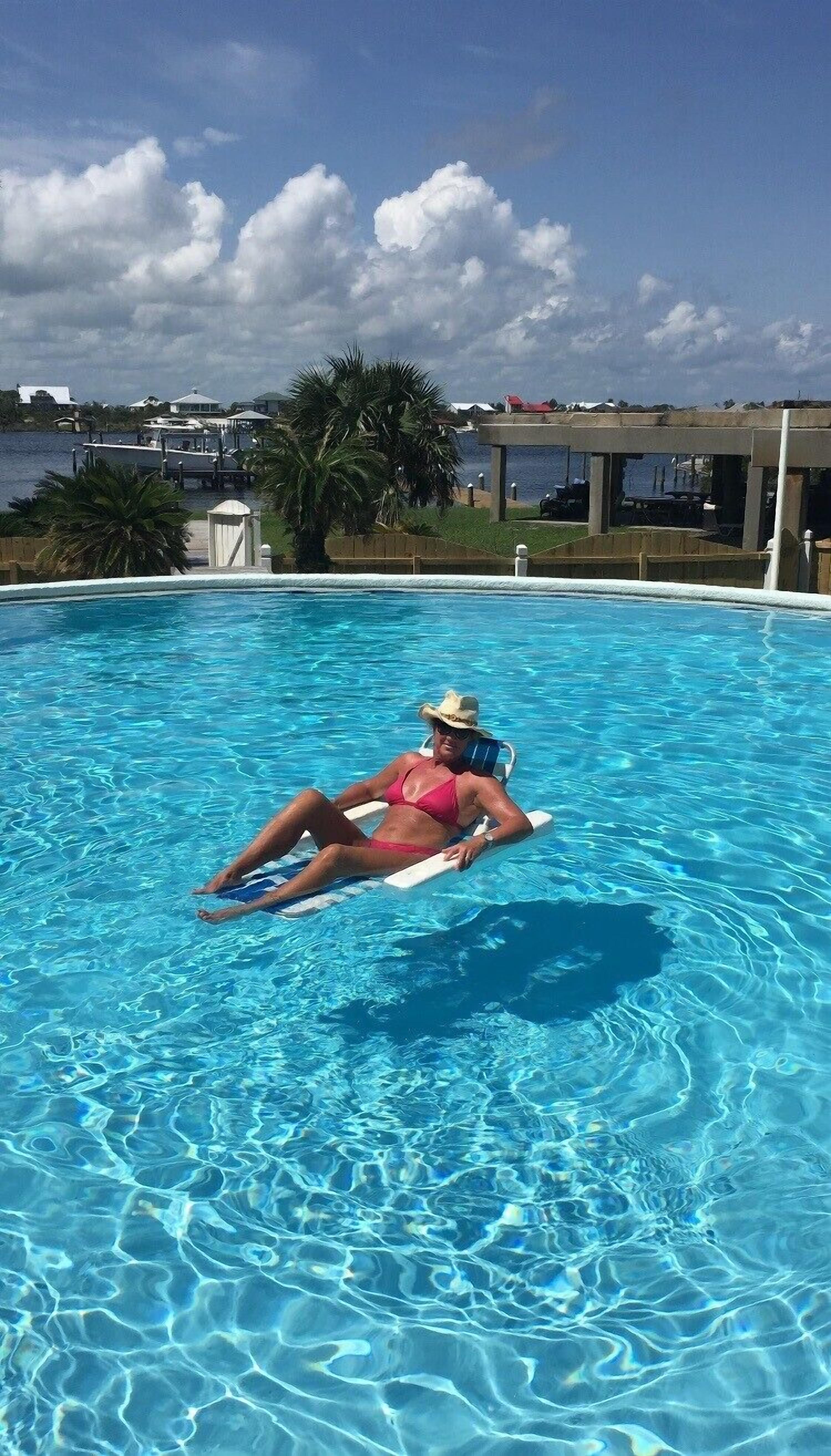A woman in a pink swimsuit lying on a lounge chair in a clear blue pool with a view of a marina and houses in the background.