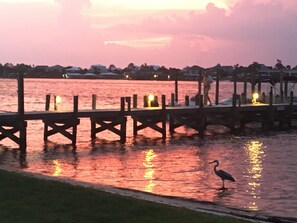 A waterfront scene with a pier extending into calm waters, a bird standing on the shore, and a sunset in the background. - Family Friendly & Quiet Riverside Condo with Pool & Fishing Piers (Pensacola)