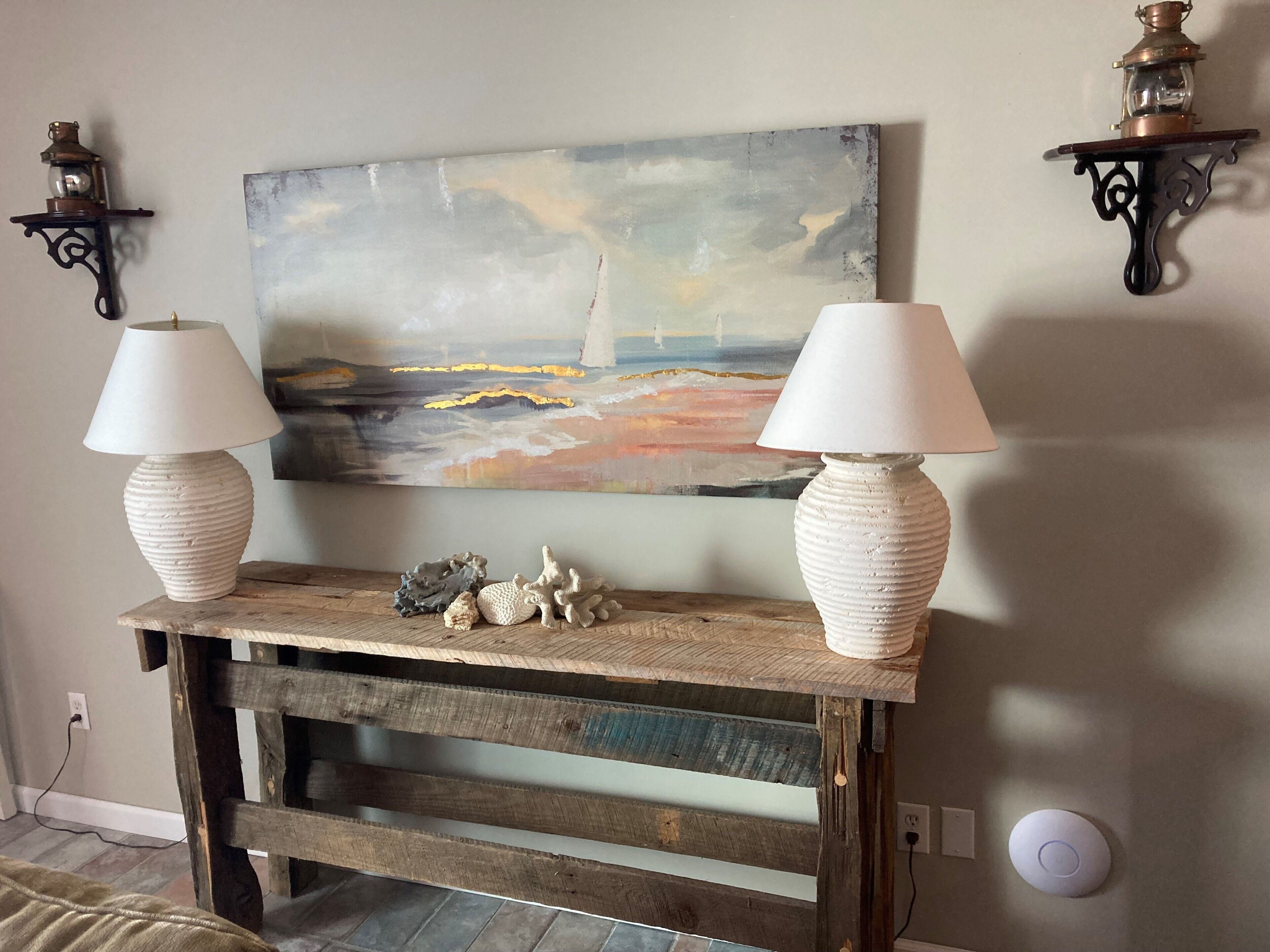A rustic wooden console table with two white lamps, a painting of a seascape, and decorative coral pieces.