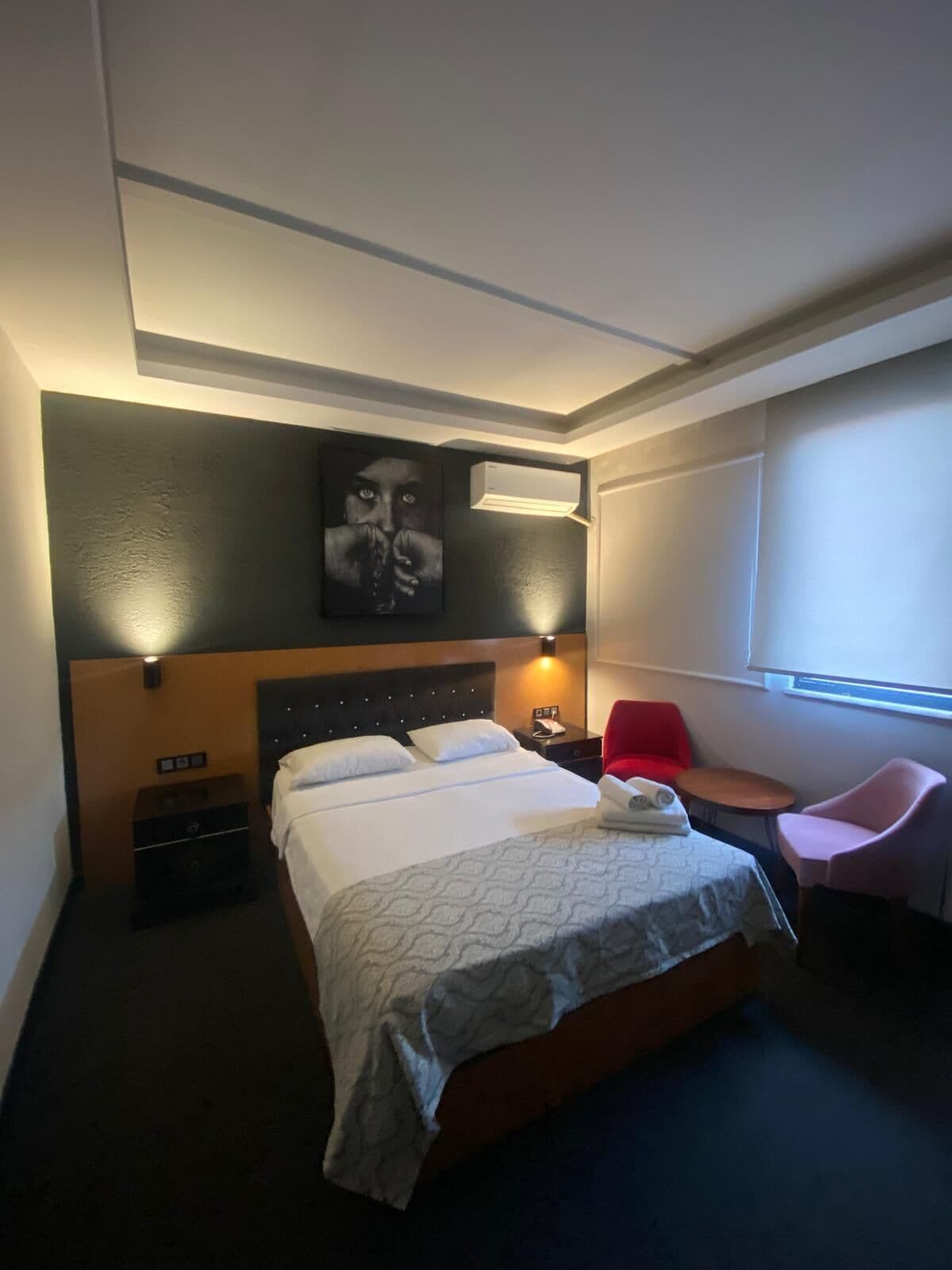 A hotel room with a bed, bedside table, chair, and a painting on the wall.