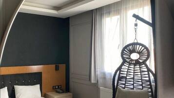 A hotel room with a bed, a hanging chair, a bedside table, and a window with curtains.