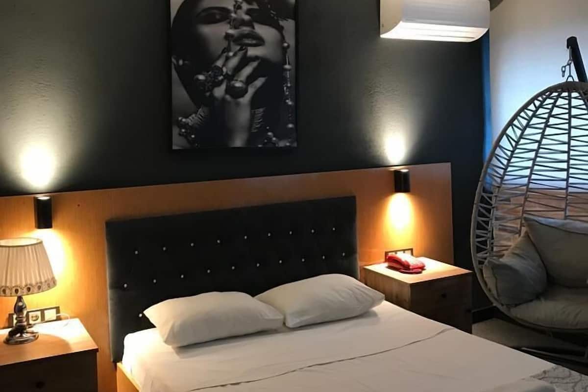 Quarto de hotel com cama, duas mesas de cabeceira com abajures, uma cadeira suspensa e um ar-condicionado fixado na parede.