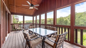 Un espace extérieur abrité, doté d’une table et de chaises à plateau en verre, d’une terrasse en bois et de grandes fenêtres offrant une vue sur la verdure environnante.