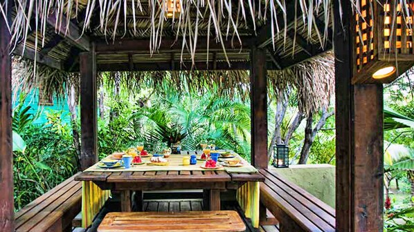 An outdoor dining area with a wooden table set for two, surrounded by lush greenery and a thatched roof structure.