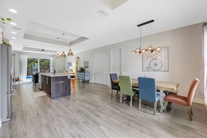 A modern kitchen with a central island, dining area with colorful chairs, and a large window providing natural light. - ISLAND LIFESTYLE FAMILY VACATION HOME SALT POOL (Naples)