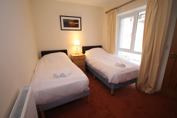 Une chambre d’hôtel avec deux lits simples, une table de chevet avec une lampe, un radiateur, une fenêtre avec des rideaux et un tableau encadré au mur.