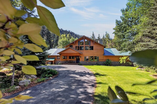 A two-story log cabin with a stone foundation, a metal roof, and a paved driveway leading to the entrance.