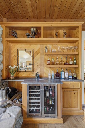 Uma área de bar em madeira com prateleiras cheias de garrafas de bebidas, copos e uma geladeira com garrafas.