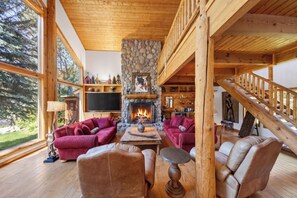 A cozy log cabin interior with a stone fireplace, wooden furniture, and a large window overlooking a forest.