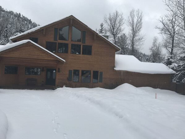 A two-story wooden cabin with a snow-covered roof and a large window on the upper floor.