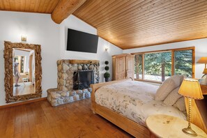 A bedroom with a stone fireplace, a bed with floral bedding, a wooden ceiling, and a large window with a view of trees.