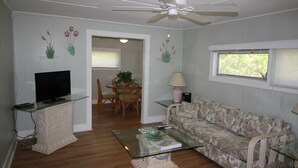 Ein Wohnzimmer mit Fernseher auf einem Glastisch, einer Couch, einem Deckenventilator und einem Fenster mit Jalousien.
