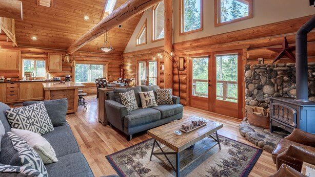 A spacious log cabin with a large living area, a stone fireplace, and a kitchen with wooden cabinets and a large window.