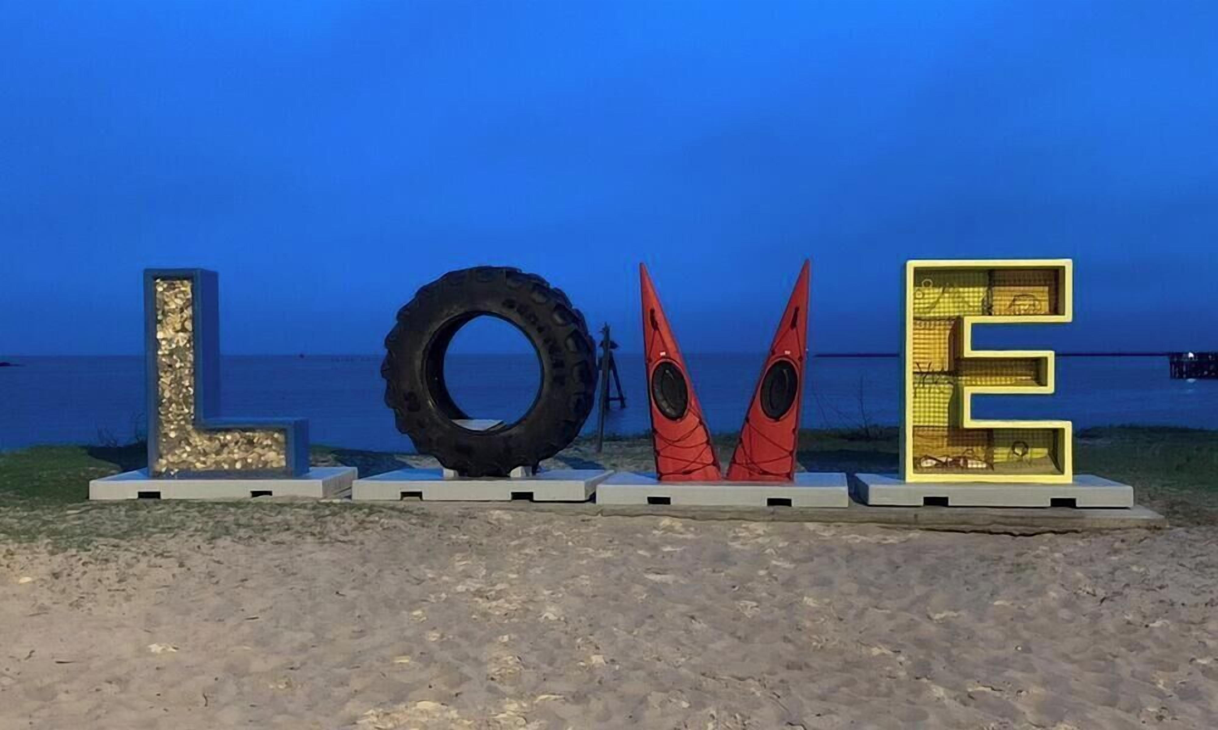 Une plage avec une grande sculpture en forme de pneu, un kayak et une structure en forme de lettre « E ».