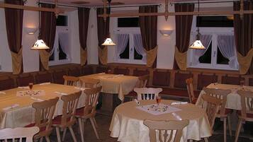 Un comedor con varias mesas y sillas, paneles de madera y ventanas grandes con cortinas.