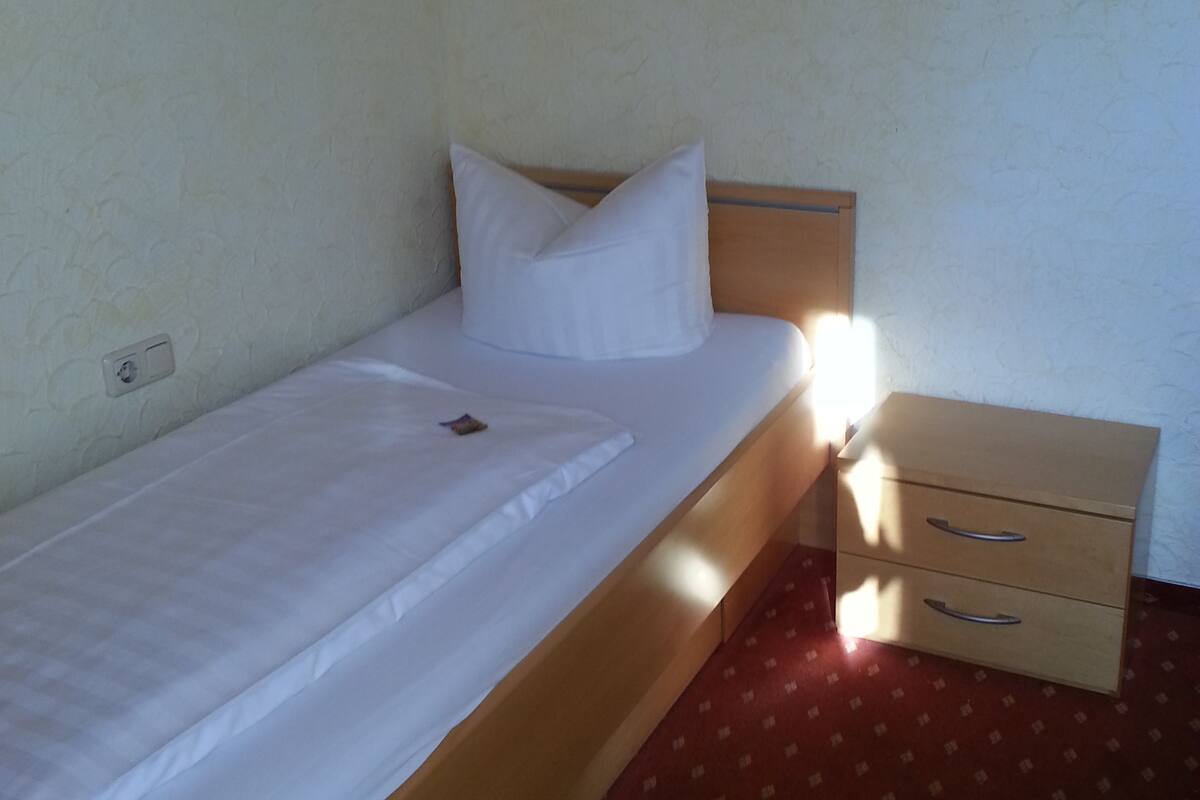 A single bed with a wooden headboard, a bedside drawer, and a patterned carpet.