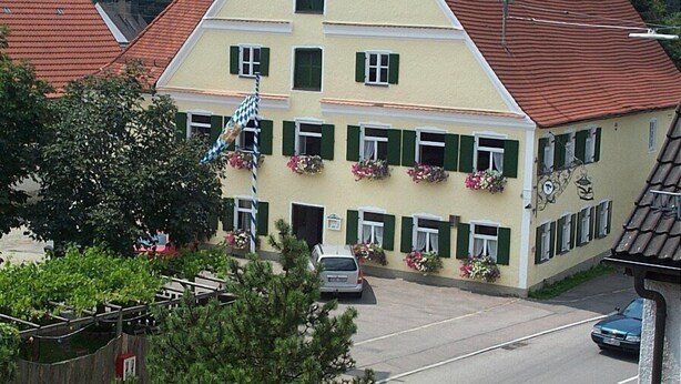 Ein mehrstöckiges Gebäude mit gelber Fassade und grünen Fensterläden, das eine zentrale Eingangstür und Blumenkästen an den Fensterbänken zeigt.