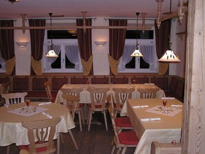 Un restaurant avec des tables et des chaises en bois, un plafond haut avec des poutres apparentes et de grandes fenêtres avec des rideaux.