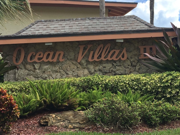 Een stenen muur met de tekst 'Ocean Villas' en een aangelegde tuin met verschillende planten en een rots.