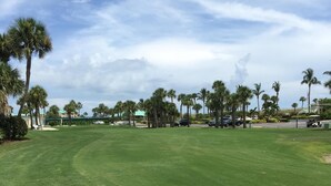 Un terrain de golf bien entretenu, avec un ciel dégagé, des palmiers et quelques voitures garées.