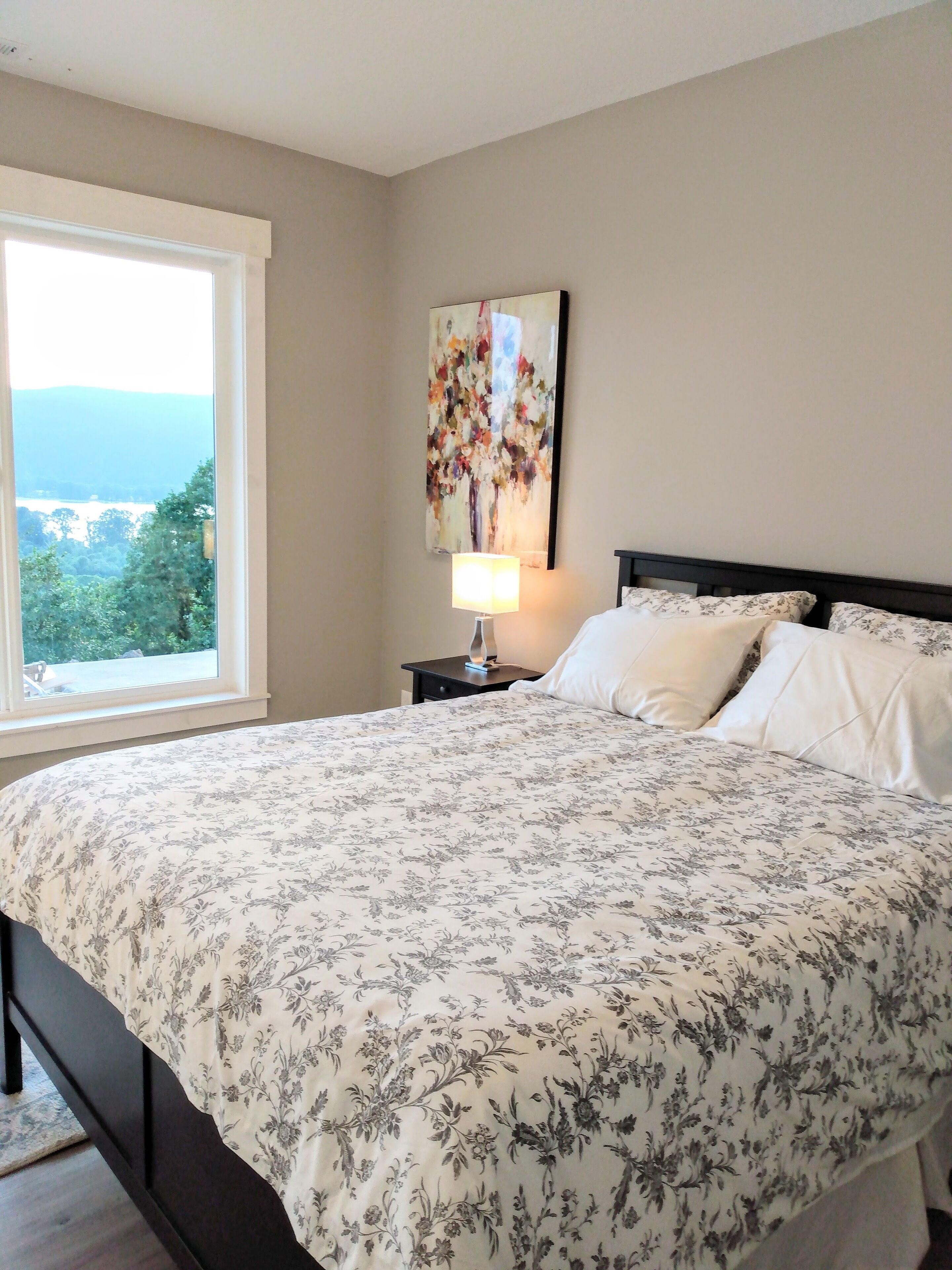A bedroom with a large bed, a nightstand with a lamp, a painting on the wall, and a window with a view of greenery.