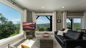 A modern living room with a large TV, comfortable seating, and a scenic view through large windows.
