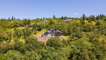 A large, modern house with a spacious driveway and multiple garages, surrounded by dense forest and other residential buildings on a hillside.