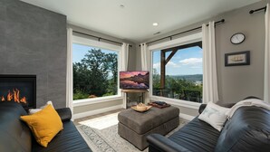 A modern living room with a fireplace, two sofas, a TV, and large windows offering scenic views.