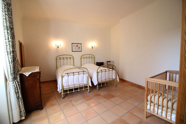 Un dormitorio con dos camas de metal, una cuna de madera, un aparador de madera y suelo de baldosas.