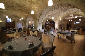 Un interior con arcos de piedra, mesas dispuestas para comer, sillas de mimbre e iluminación colgante.
