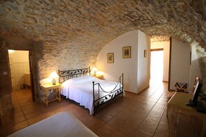 A bedroom with a stone ceiling, a bed with white linens, a nightstand with a lamp, and a wooden chair. - La Bastide du Vebron (Grospierres)