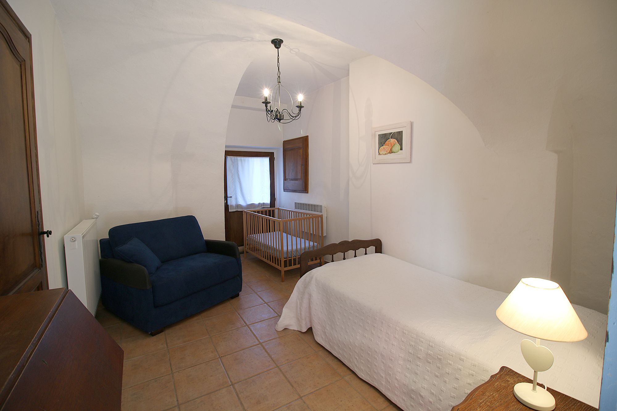 A room with a crib, a bed, a blue chair, a wooden table, and a lamp.