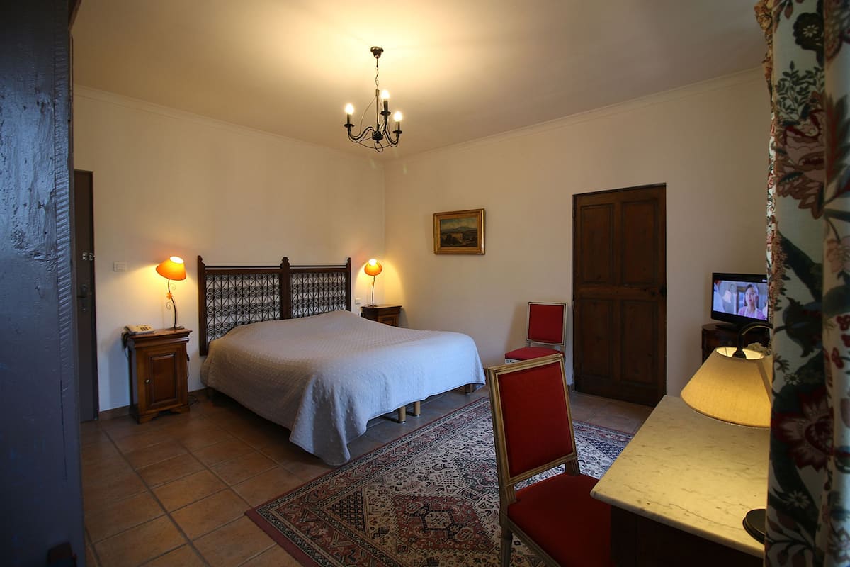Un dormitorio con una cama, dos lámparas de noche, un televisor, un escritorio con lámpara, una silla, una alfombra y una puerta.