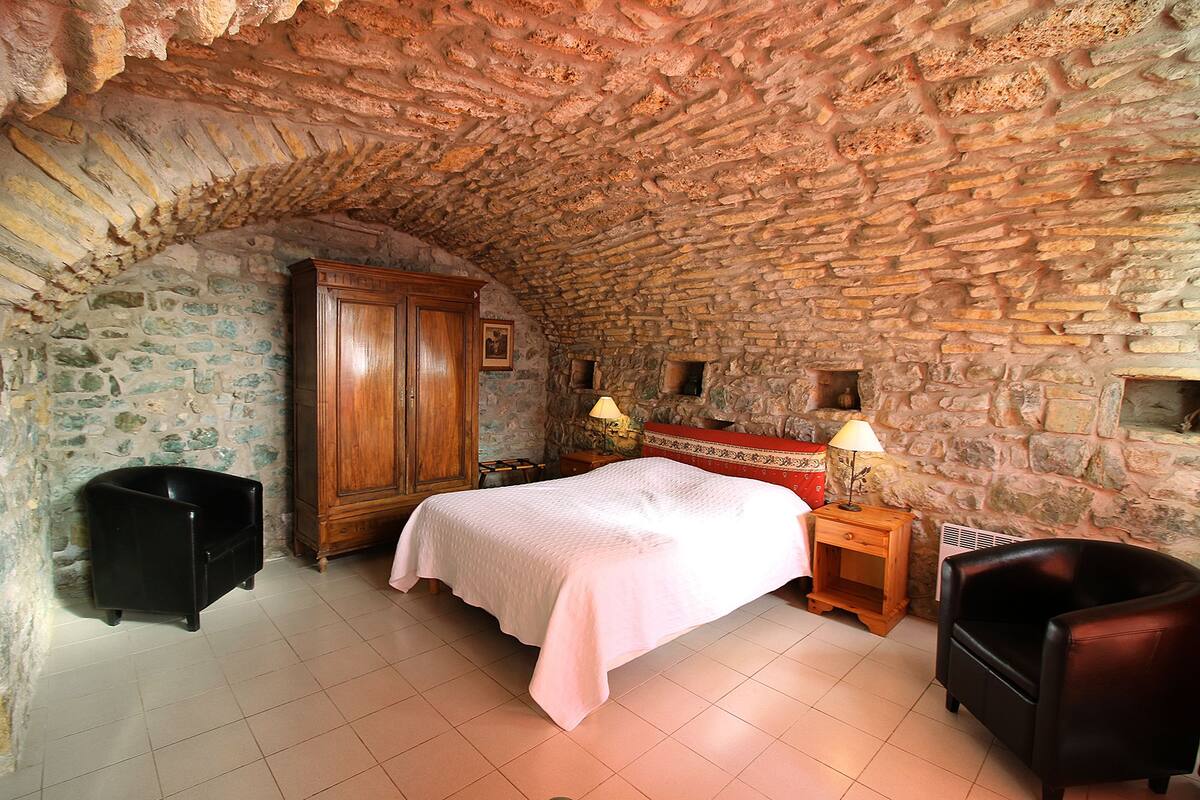 Un dormitorio con techo de piedra, un armario de madera, una cama con cubrecama blanco, dos mesitas de noche con lámparas y un sillón de cuero.