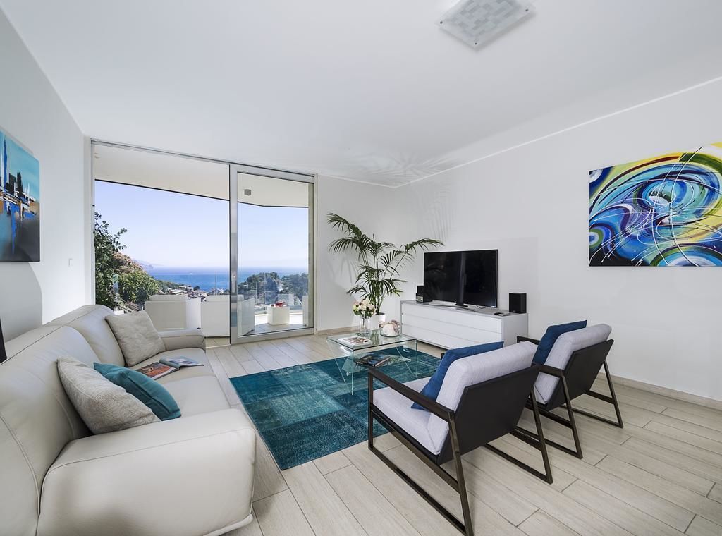 A modern living room with a sofa, two armchairs, a coffee table, and a TV. There is a large window offering a view of the sea.