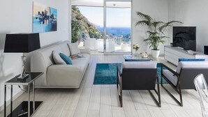 A modern living room with a sofa, coffee table, TV, and a large window offering a city view. - Isola Bella Infinity Suites (Taormina)