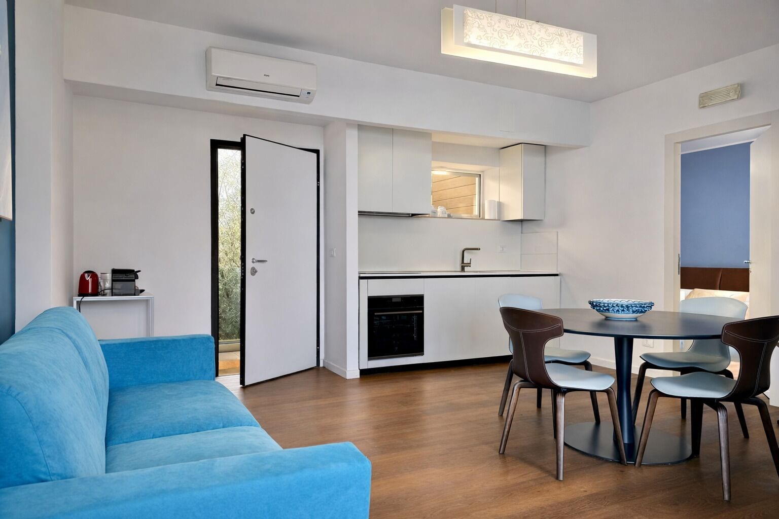 A modern living room with a blue sofa, a dining area with a table and chairs, and a kitchen with white cabinets and a built-in oven.