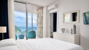 A bedroom with a large bed, a dresser, a mirror, and a TV. There is a view of the ocean through a sliding glass door. - Isola Bella Infinity Suites (Taormina)