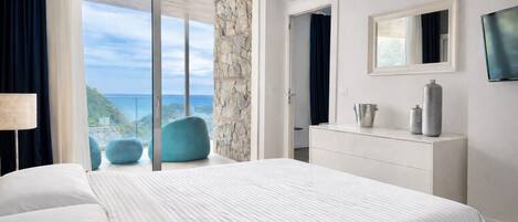 A bedroom with a large bed, a dresser, a mirror, and a TV. There is a view of the ocean through a sliding glass door.